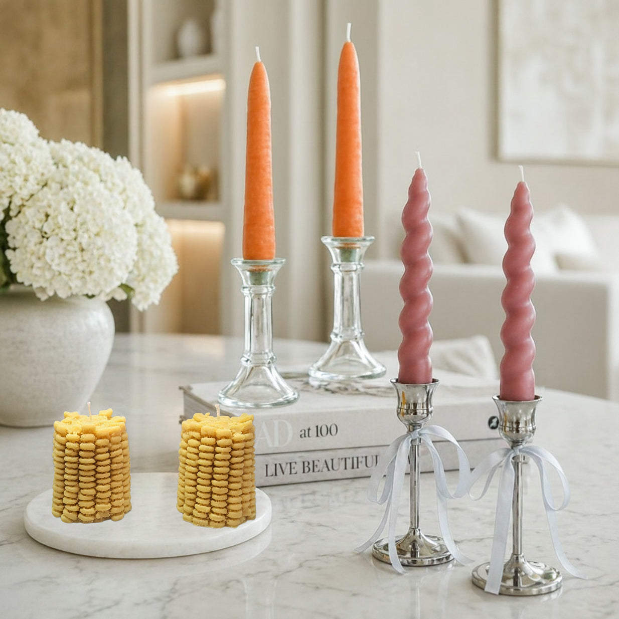 Luxury soy wax candle display on white marble featuring pairs of corn, carrot and twisted taper candles at varied heights with glass and metal holders, styled in a soft, elegant interior setting