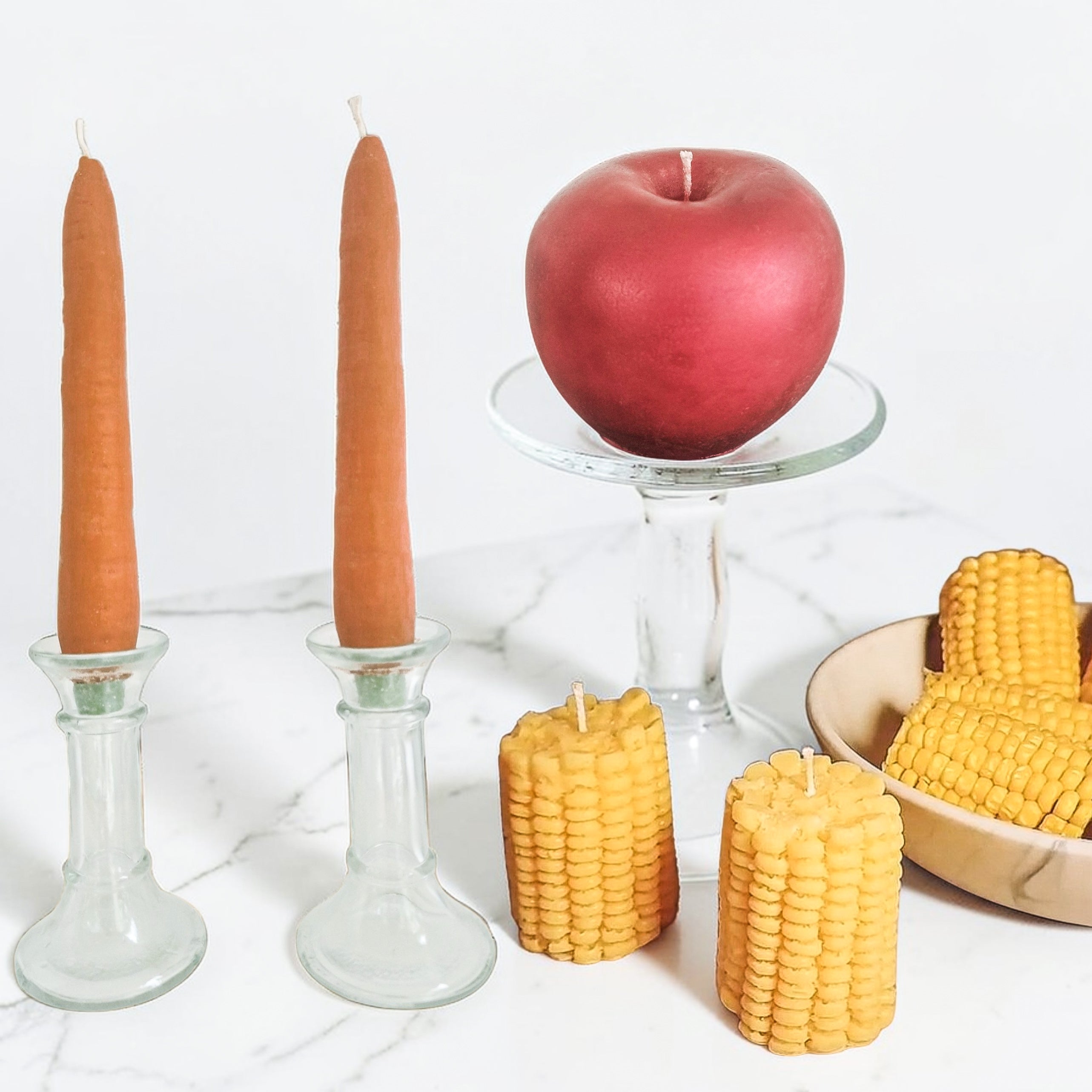 Handcrafted carrot and corn shaped soy wax candles displayed in glass holders beside a red apple on a pedestal, styled on a marble surface.