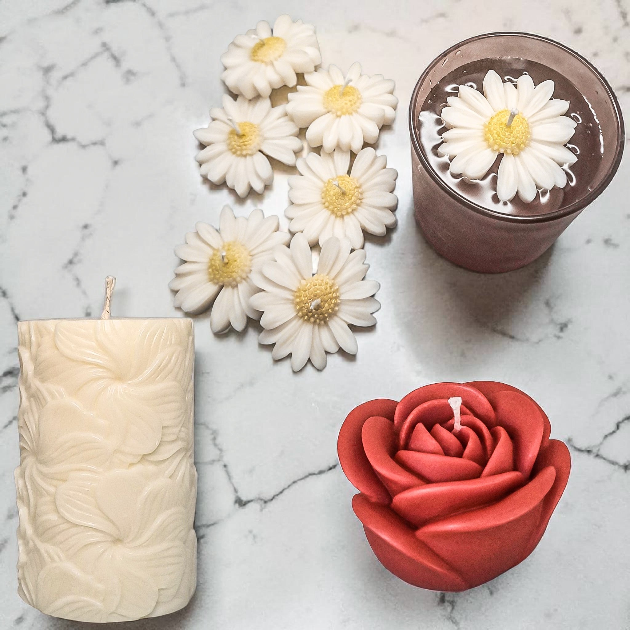 Hand poured soy wax decorative candles including a white floral candle, a red rose candle, and a glass with a white daisy flower candles on a marble surface.
