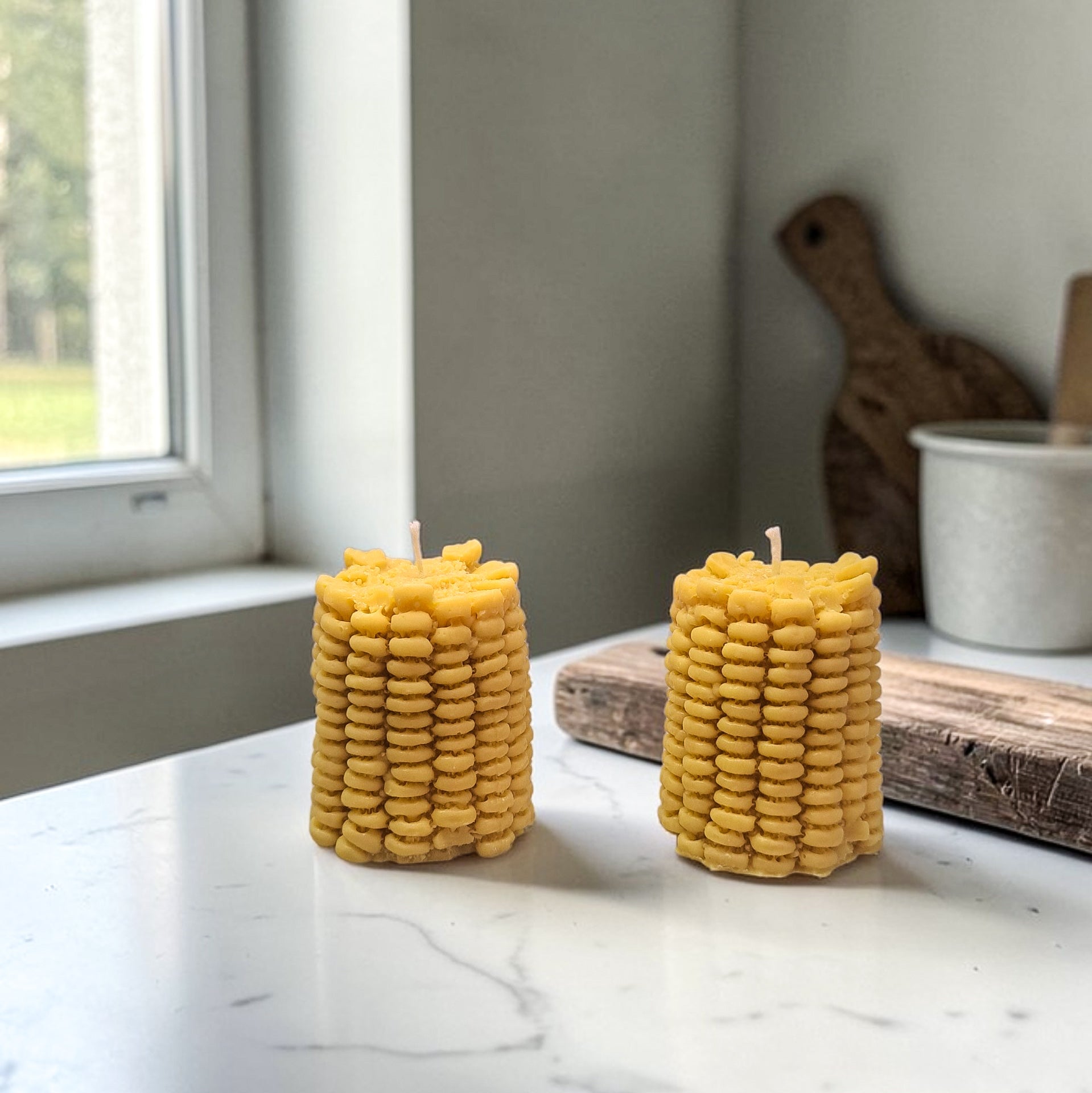 Two yellow corn on the cob soy wax candles on a marble surface with a window in the background.