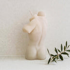 Ceramic sculpture of a nude figure on a white surface with a plant in the foreground