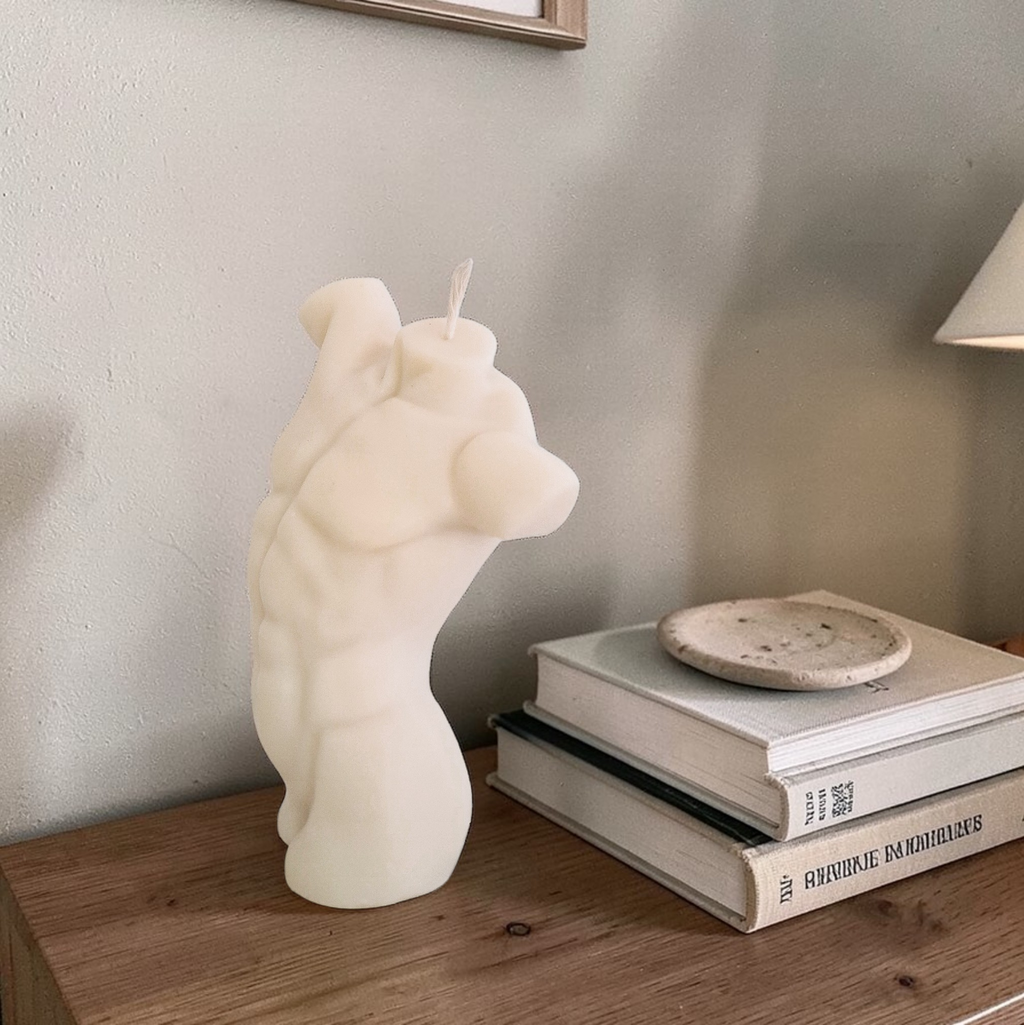 Muscular white male torso-shaped soy wax candle on a wooden surface with books and a small dish in the background.