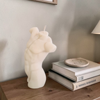Muscular white male torso-shaped soy wax candle on a wooden surface with books and a small dish in the background.