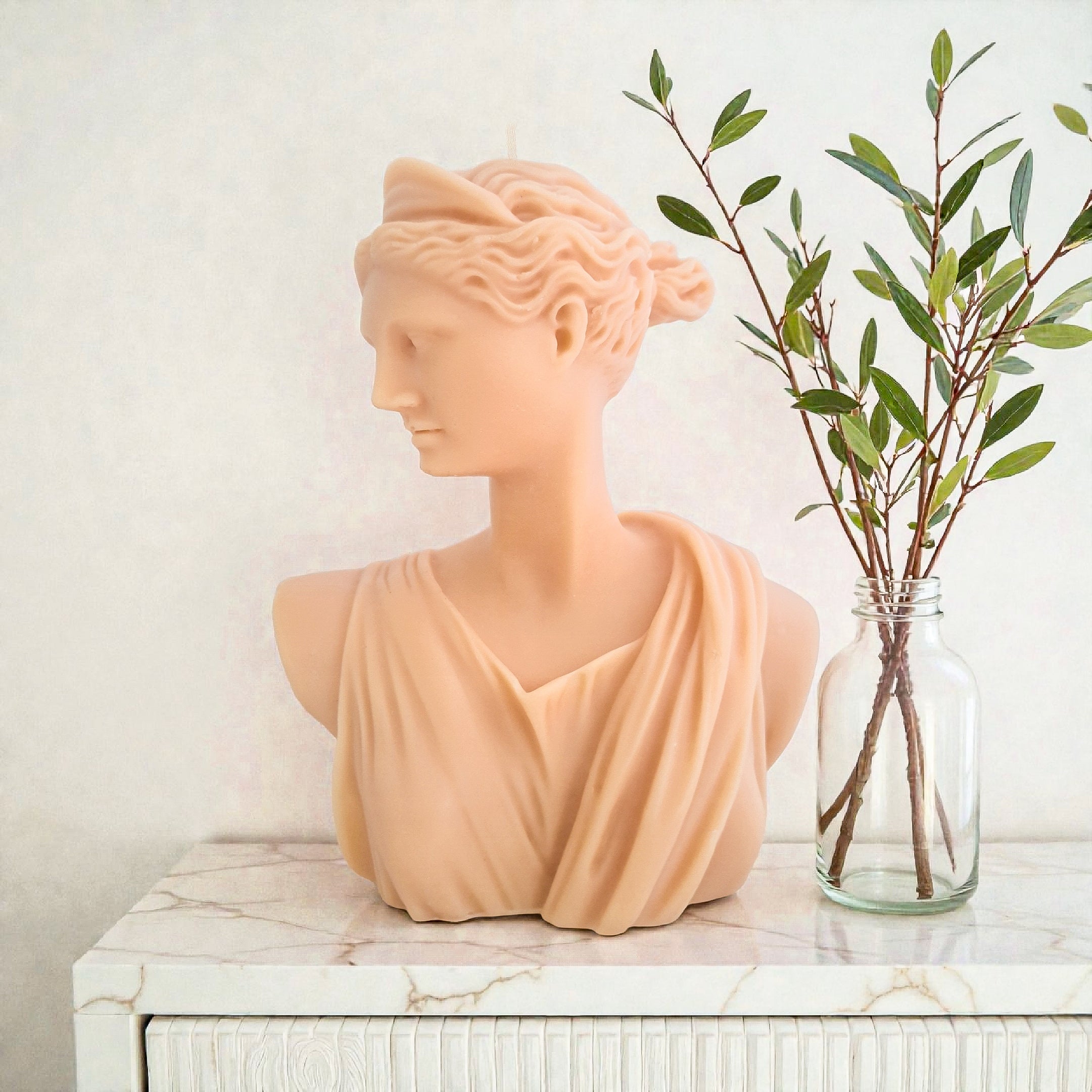 Artemis Bust soy wax sculpture e on a marble surface with a vase of greenery.
