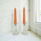 Two orange candles in glass candlesticks on a white surface with a tiled wall and window in the background.