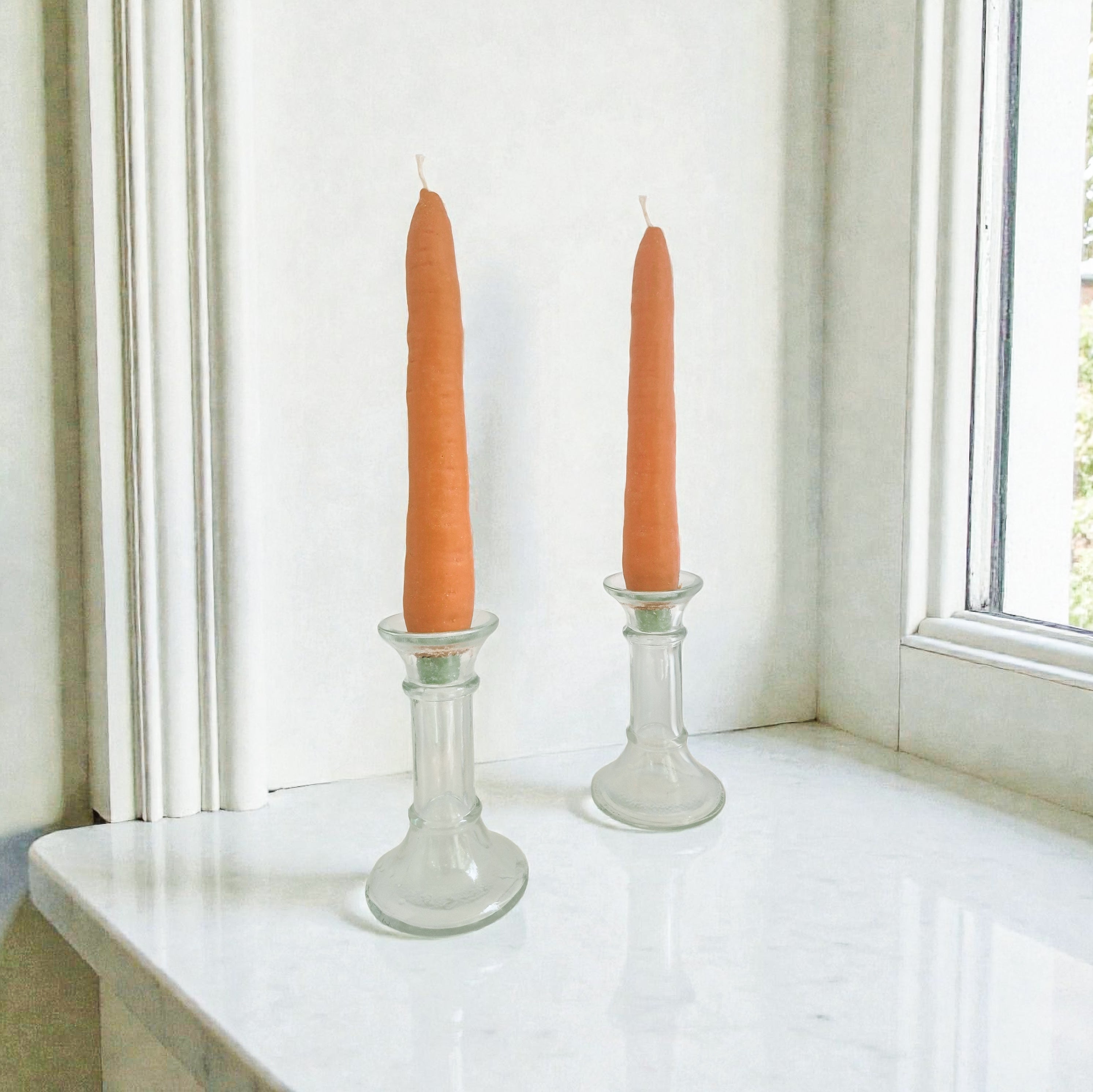 Two orange candles in glass candlesticks on a white surface near a window.