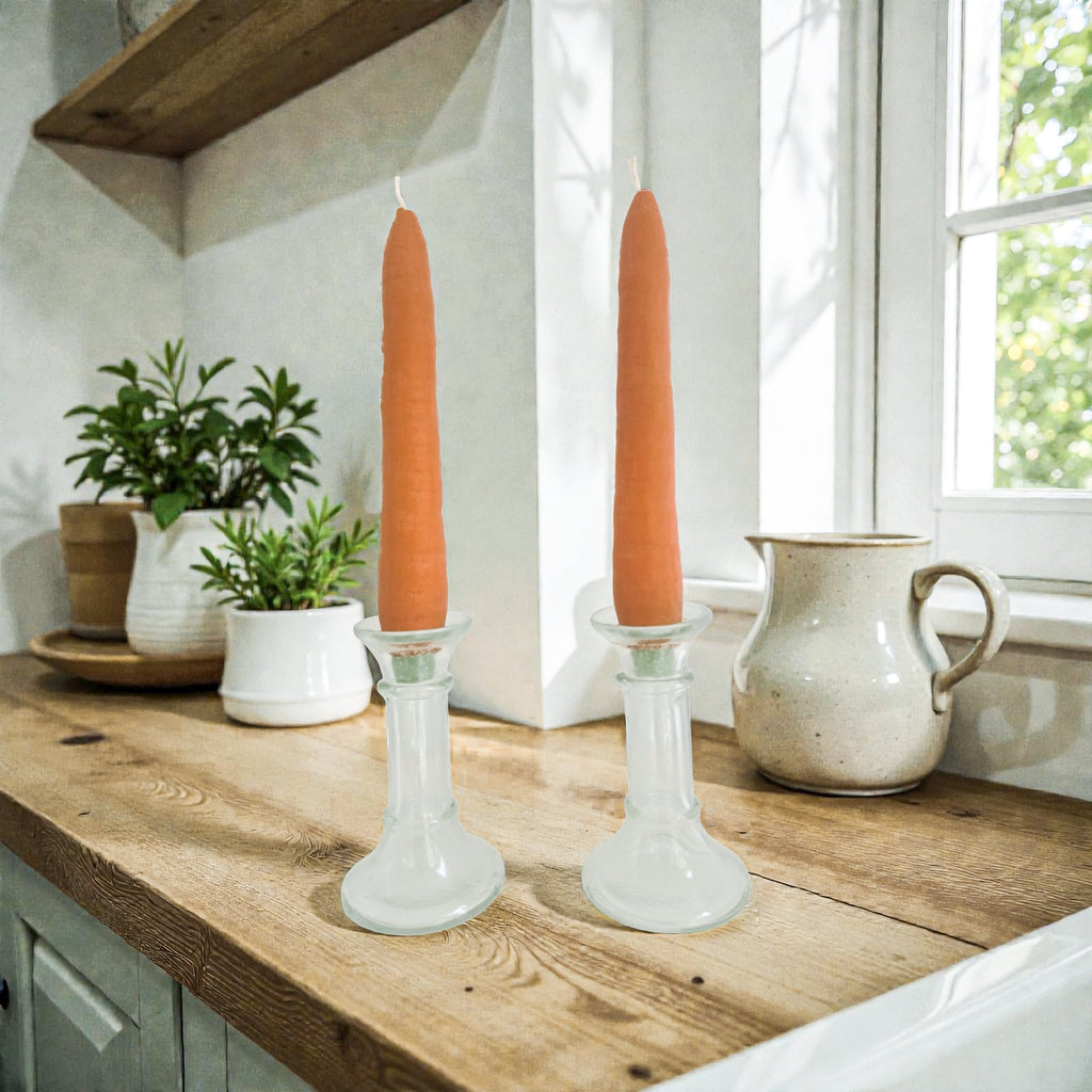 Two orange candles in glass candlesticks on a wooden surface with a window in the background.