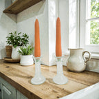 Two orange candles in glass candlesticks on a wooden surface with a window in the background.