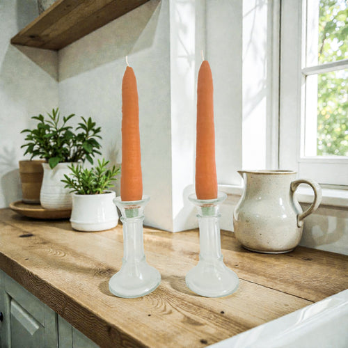 Two orange candles in glass candlesticks on a wooden surface with a window in the background.
