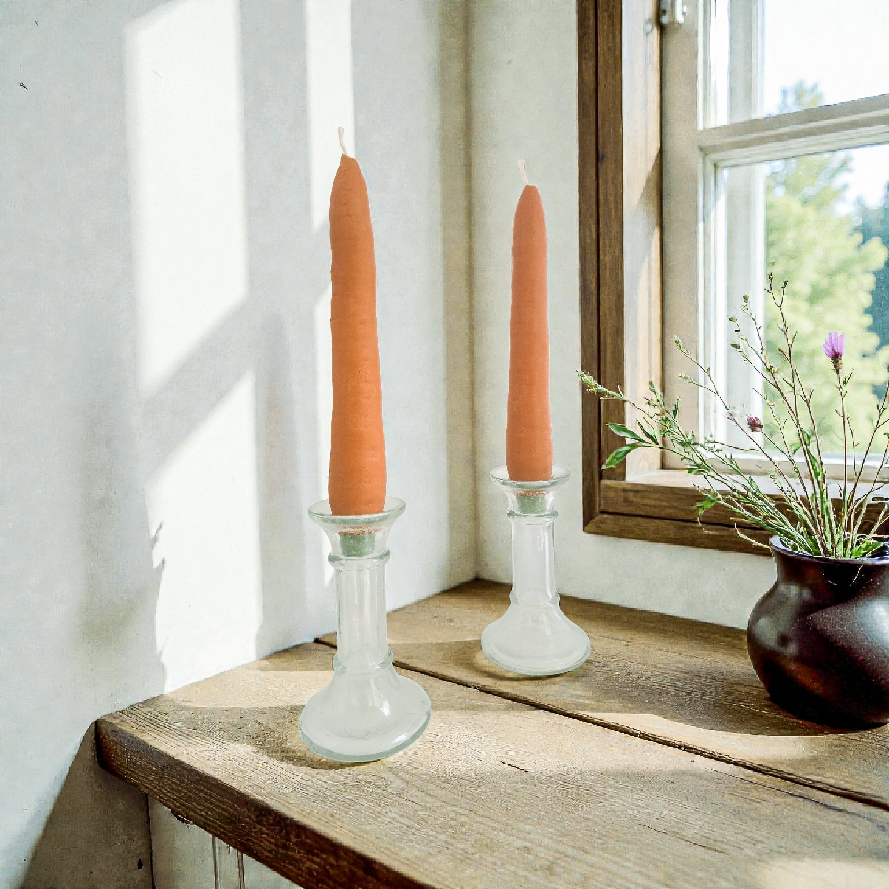 Two orange candles in glass candlesticks on a wooden surface with a window in the background.