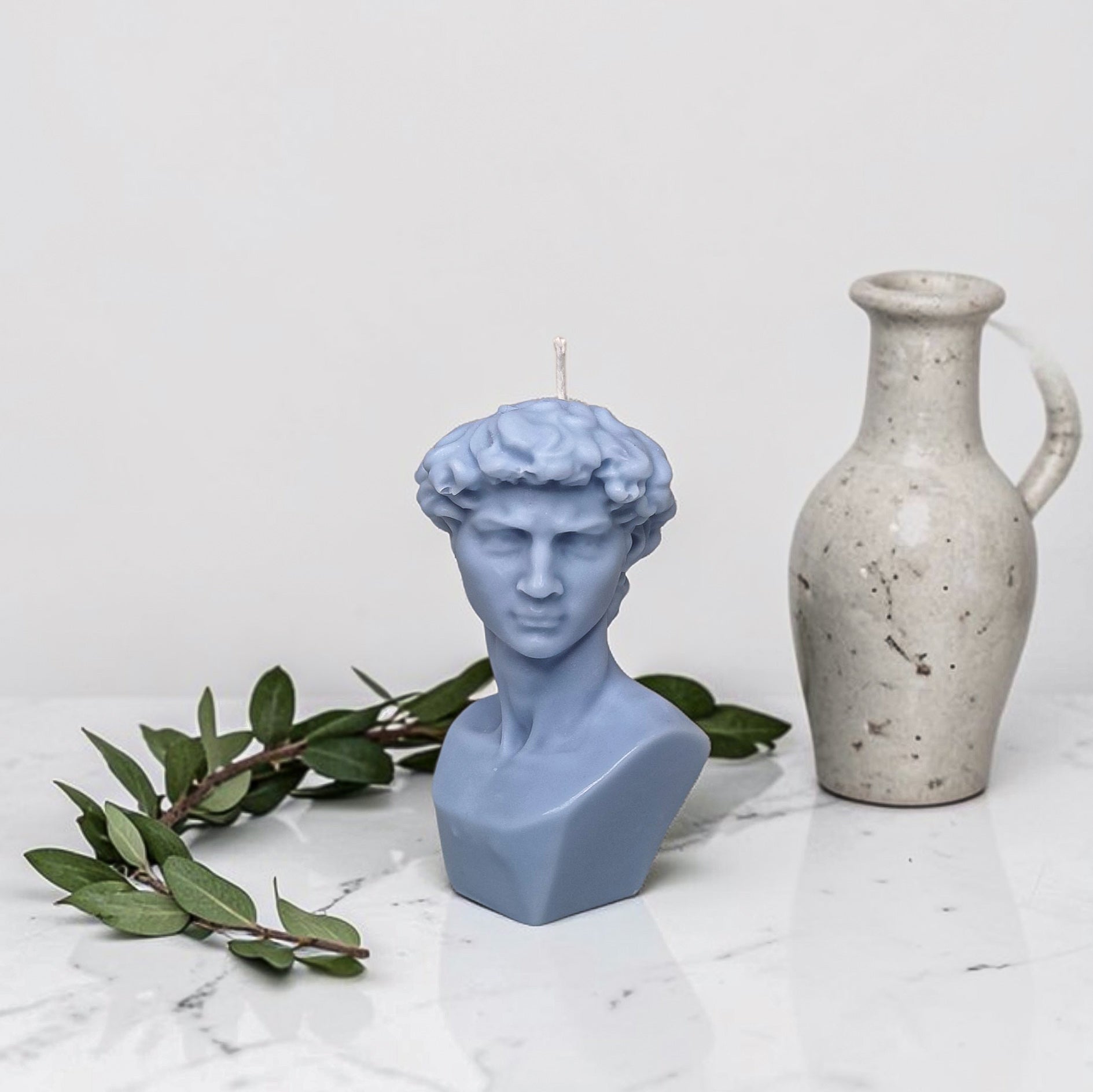 Blue bust-shaped soy wax candle with a white vase and green leaves on a light gray background