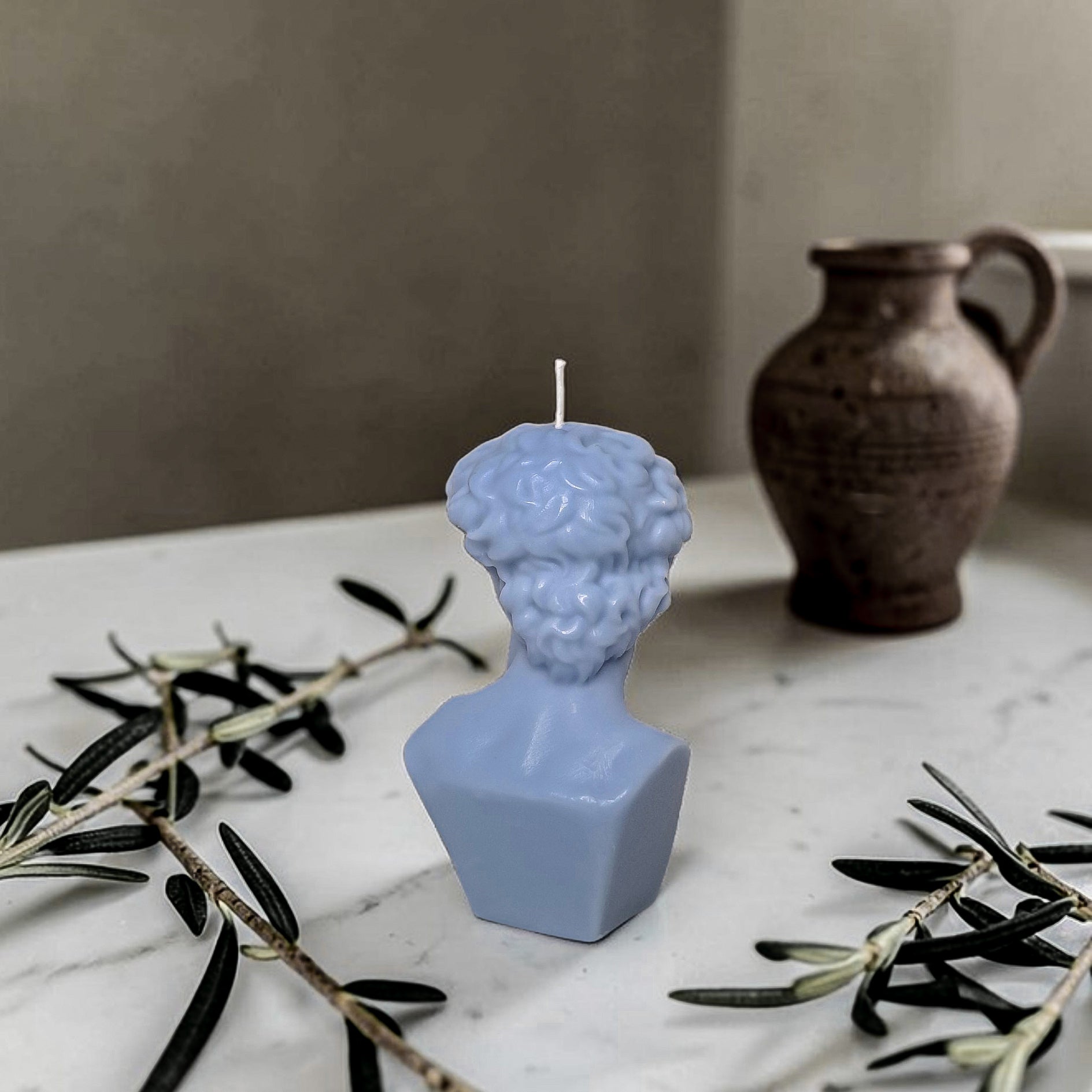 Blue bust-shaped soy wax candle on a marble surface with olive branches and a brown vase in the background.