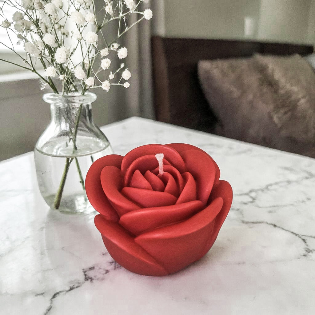 Hand poured soy wax red rose-shaped candle on a marble surface with a vase of white flowers in the background.