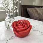 Hand poured soy wax red rose-shaped candle on a marble surface with a vase of white flowers in the background.