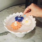 Hand lighting colourful daisy soy wax candles in a decorative bowl on a marble surface.