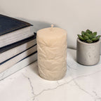 Beige textured floral pattern pillar soy wax candle on a marble surface with books and a succulent in the background