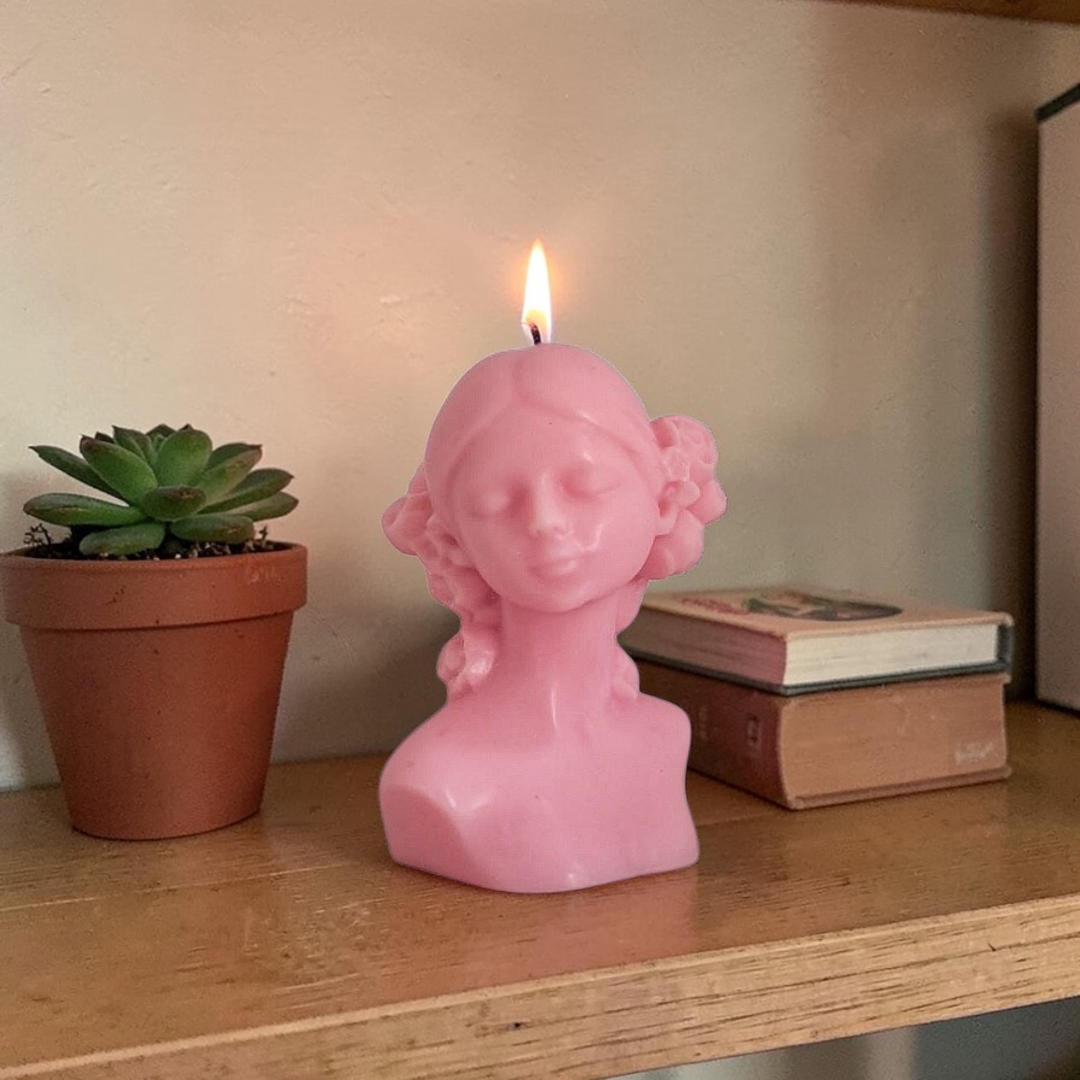 Pink bust-shaped soy wax candle with a flame on a wooden surface next to a potted plant and books.