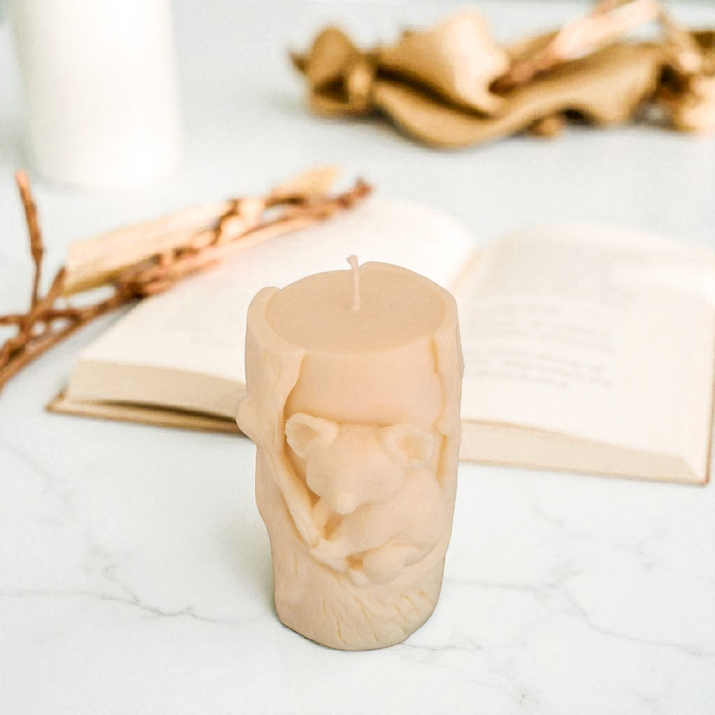 Beige soy wax candle shaped like a tree stump with a koala bear on a white surface with gold branches in the background