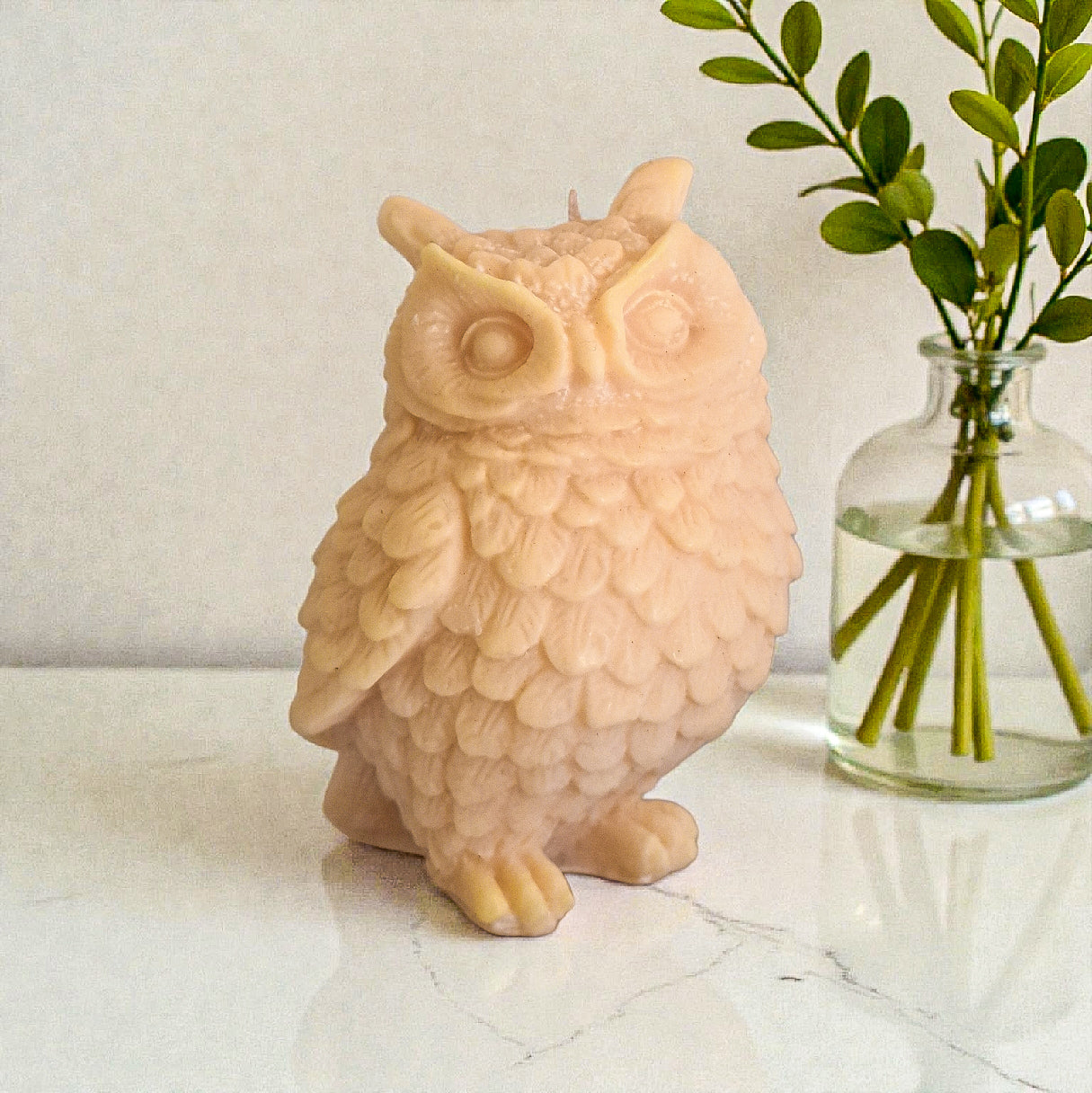 Owl-shaped candle on a marble surface with a vase of greenery in the background