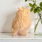 Owl-shaped candle on a surface with a vase of greenery in the background