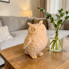 Owl-shaped soy wax candle on a wooden table with a vase of greenery in a living room setting.