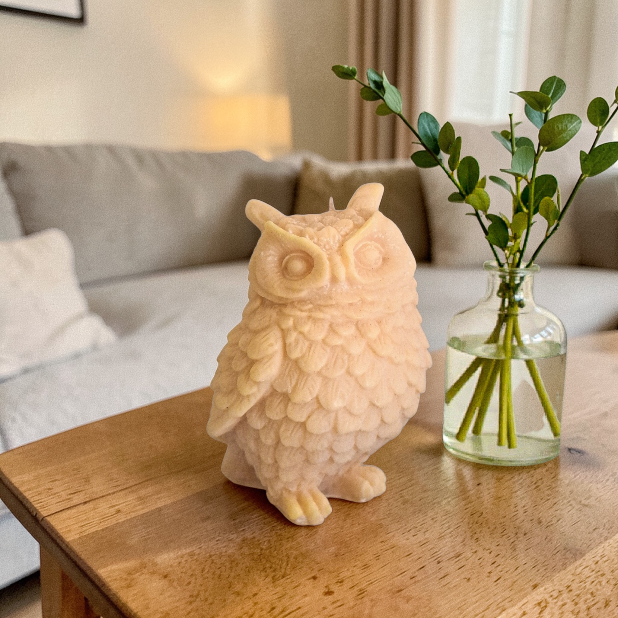 Owl-shaped soy wax candle on a wooden table with a vase of greenery in a living room setting.