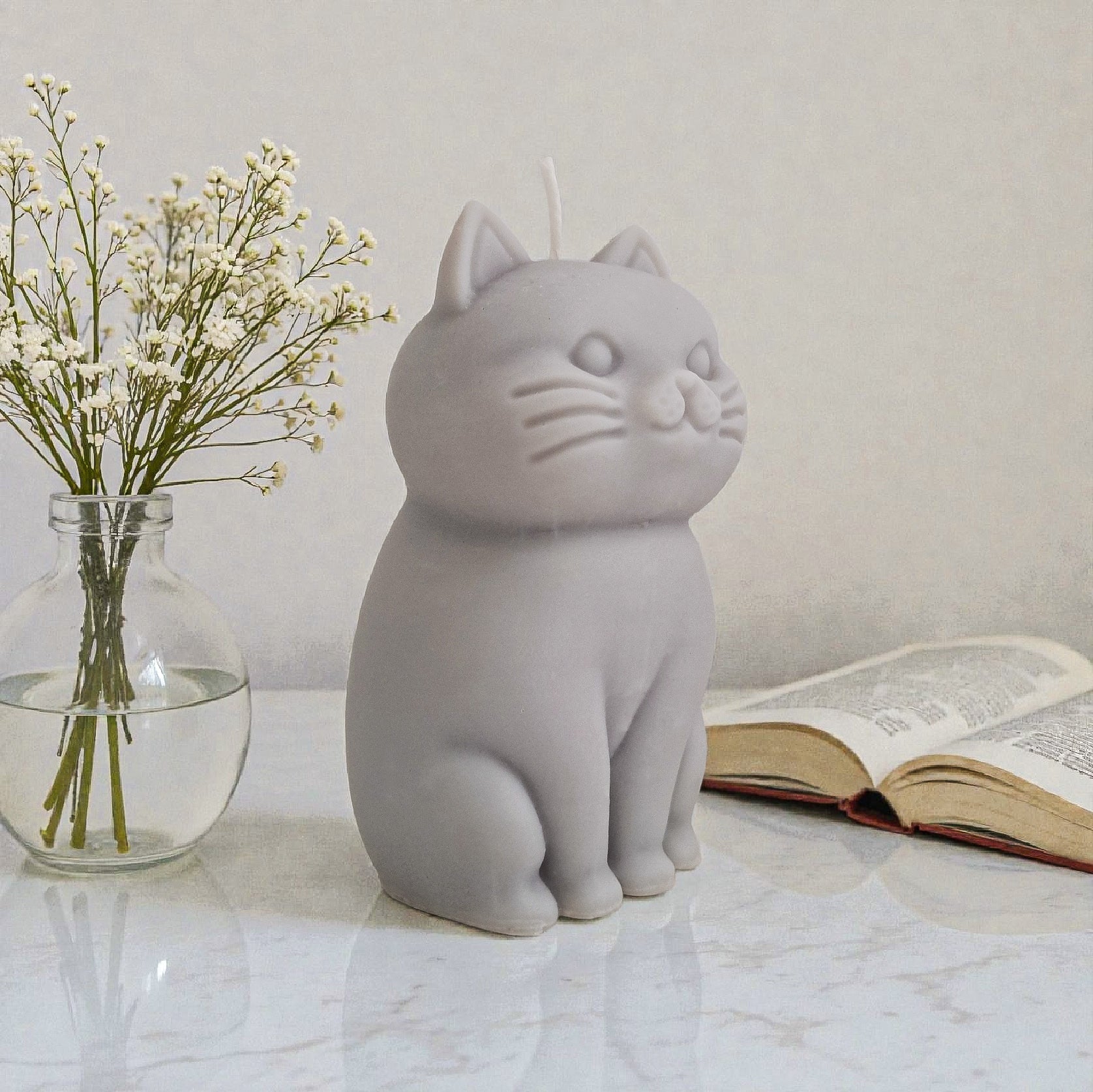 Sculptural cat-shaped soy pillar candle styled on marble surface with floral vase flowers and an open book in the background.