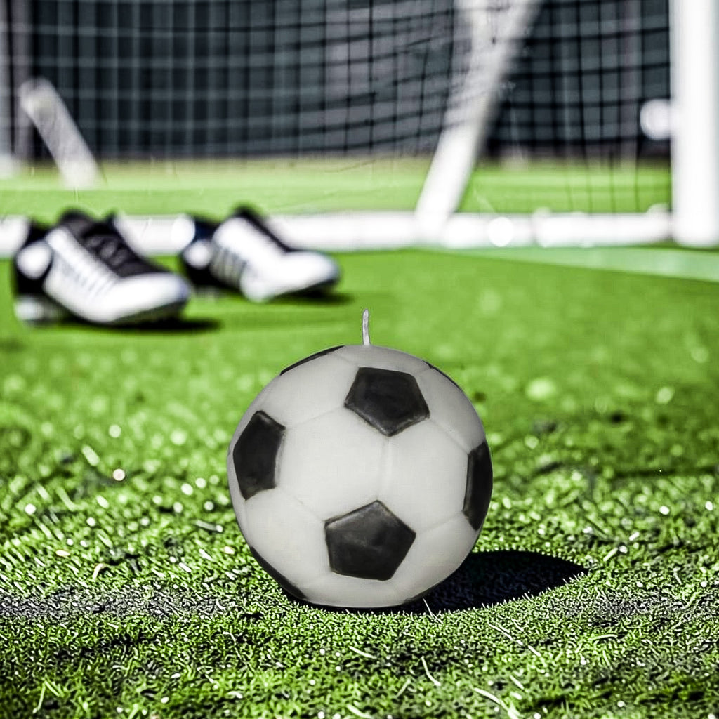 Classic black and white soy-wax candle soccer ball on a green field with goal post in the background and soccer shoes