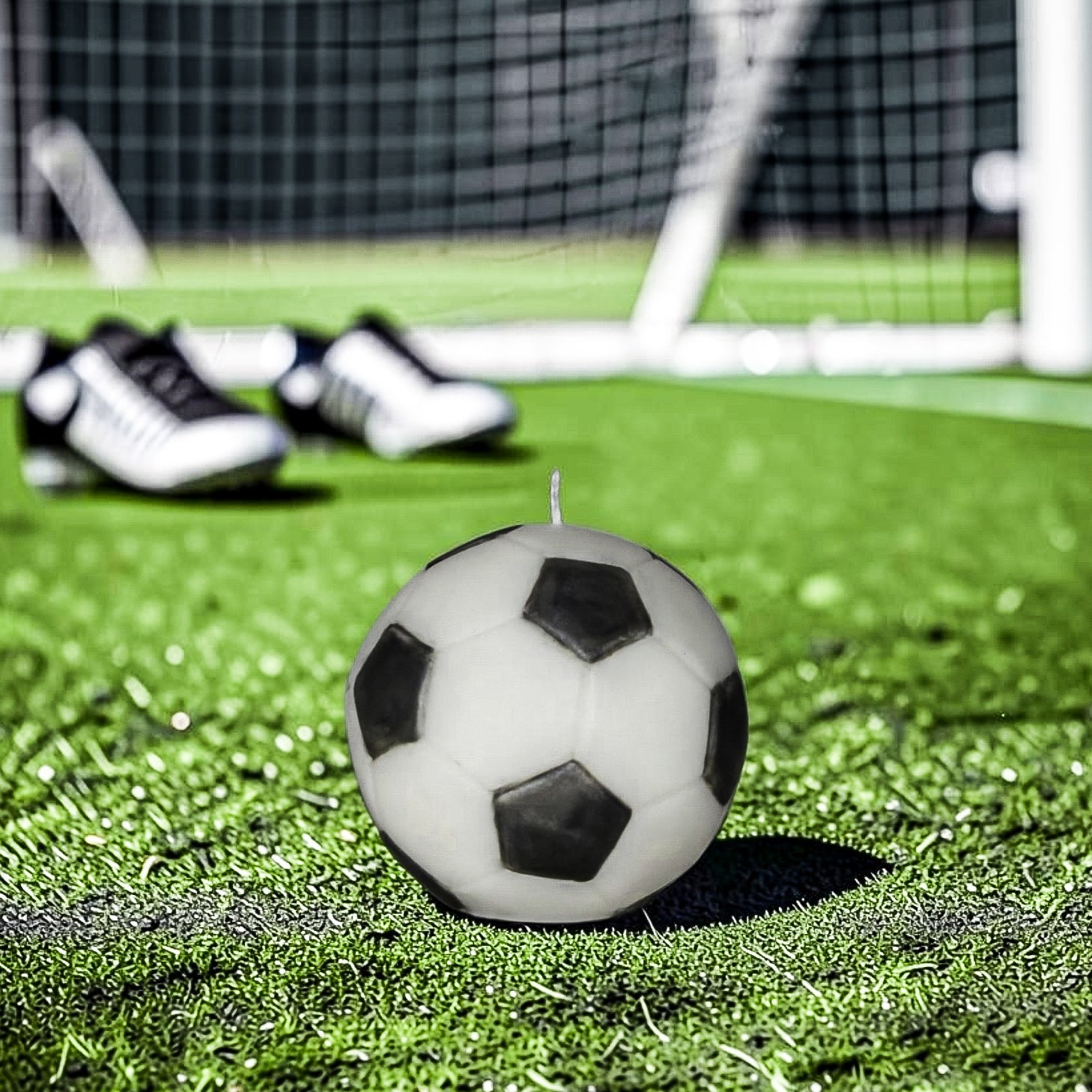 Classic black and white soy-wax candle soccer ball on a green field with goal post in the background and soccer shoes