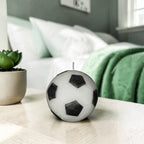 Classic black and white soy-wax candle soccer ball on a a desk in a bedroom with a plant on the side and a bed in the background