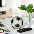 Classic black and white soy-wax candle soccer ball on a a marble table in a TV room with TV remote on the side and a plant in the background.