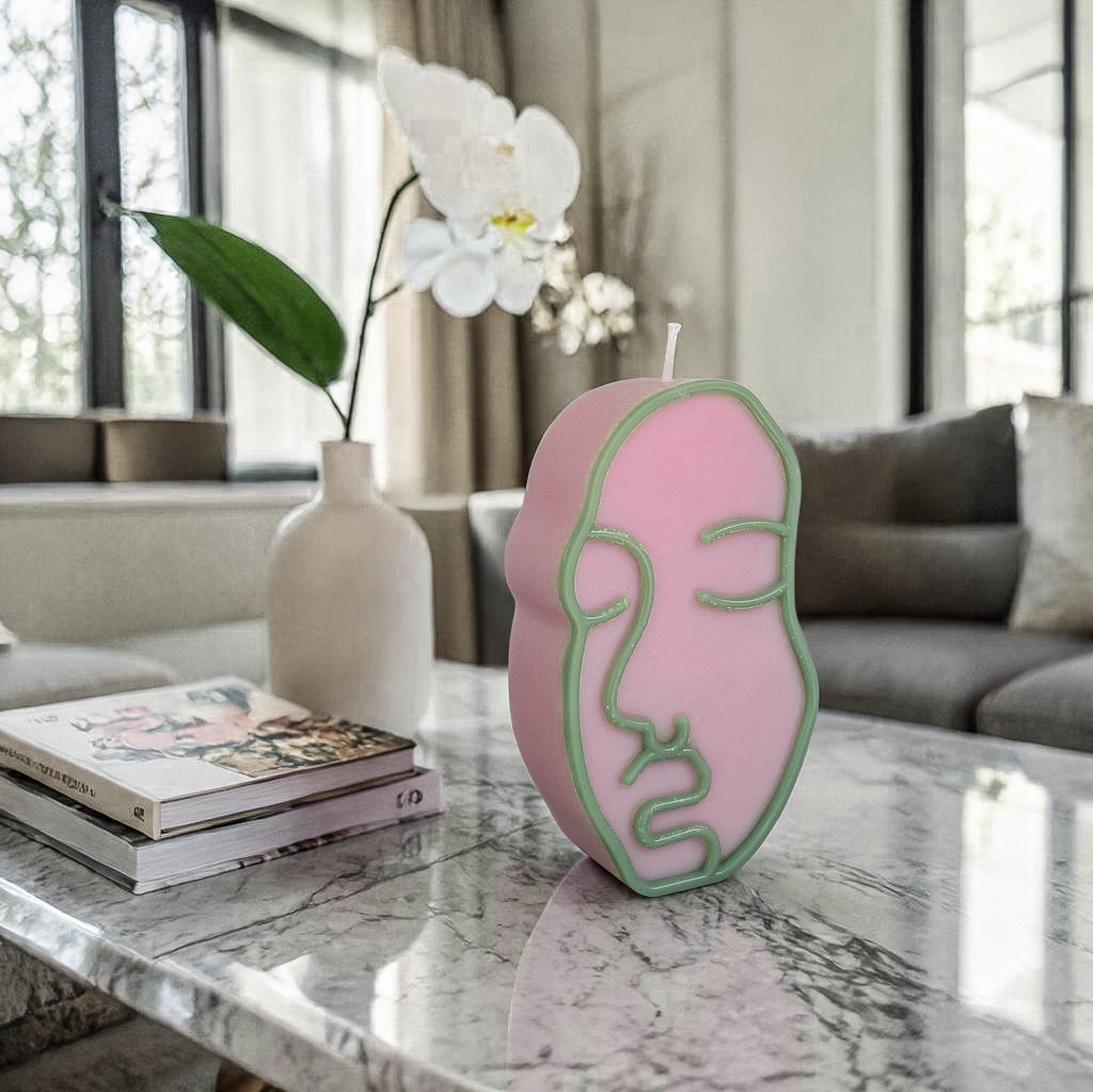 Pink face-shaped hand poured soy wax candle on a marble table with a vase and books in the background