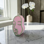 Pink and green abstract face-shaped  hand poured soy wax candle on a marble surface with a vase of white flowers in the background.