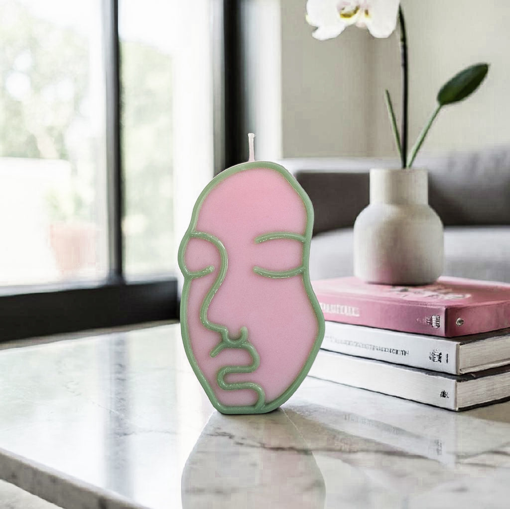 Pink and green face-shaped hand poured soy wax candle on a marble surface with a vase and books in the background.