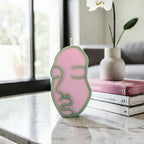 Pink and green face-shaped hand poured soy wax candle on a marble surface with a vase and books in the background.