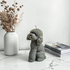 Gray dog-shaped soy wax candle on a marble surface with a vase of dried flowers and books in the background.