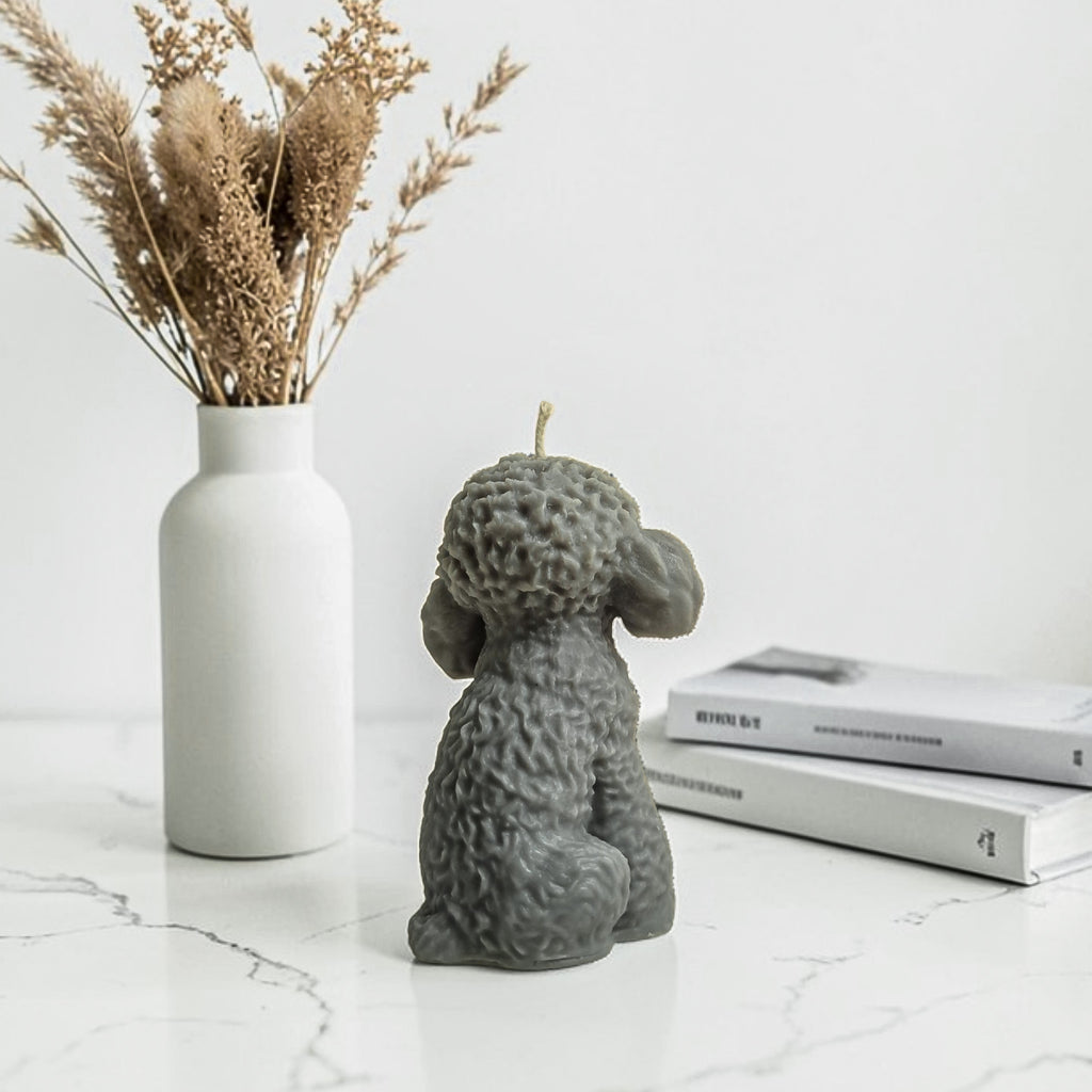 Gray dog-shaped soy wax candle on a marble surface with a vase of dried plants and books in the background.
