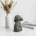 Gray dog-shaped soy wax candle on a marble surface with a vase of dried plants and books in the background.