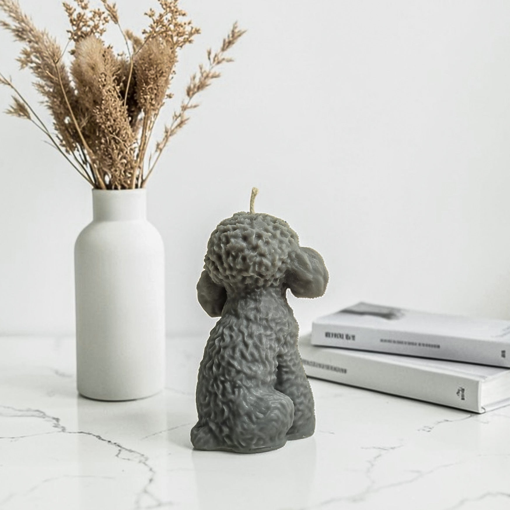 Gray dog-shaped soy wax candle on a marble surface with a vase of dried plants and books in the background.