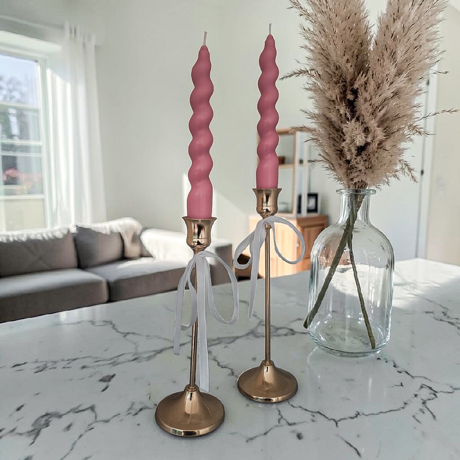 Hand poured soy wax twisted pink candles on gold candlesticks with a vase of dried pampas grass on a marble surface.