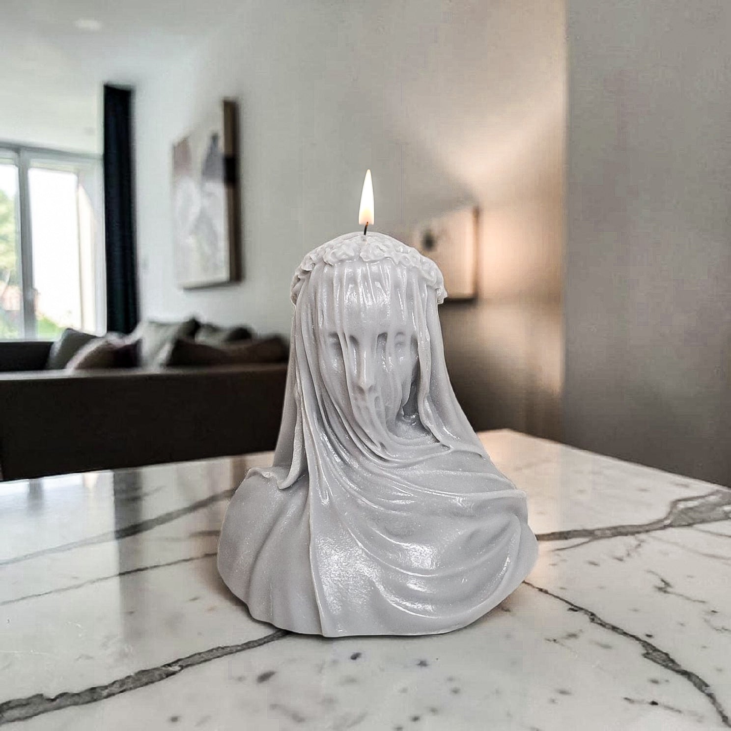 Hand poured soy wax Candle in the shape of a draped figure on a marble surface with a blurred indoor background