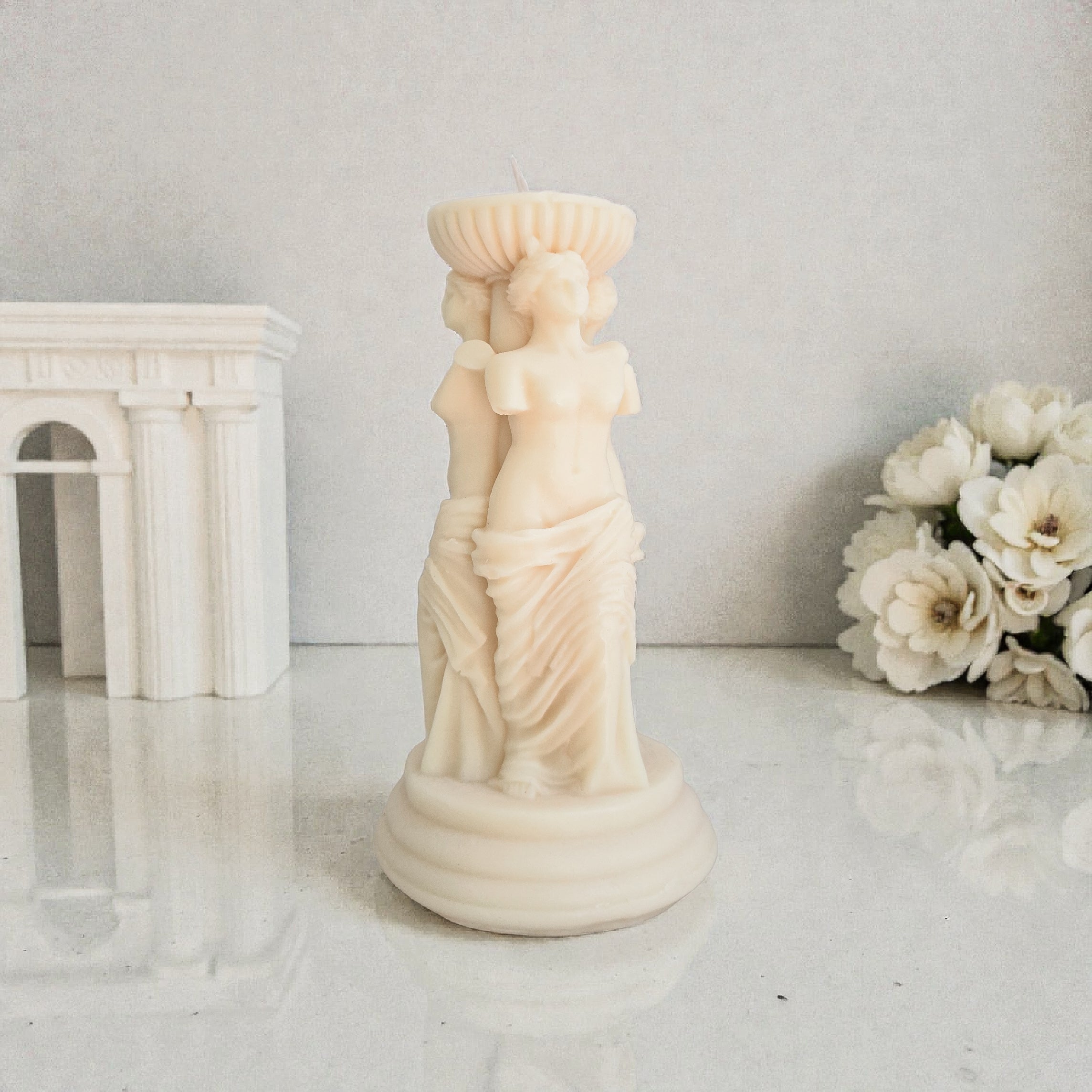 Decorative statue of a classical figure on a white surface with flowers and architectural model in the background.