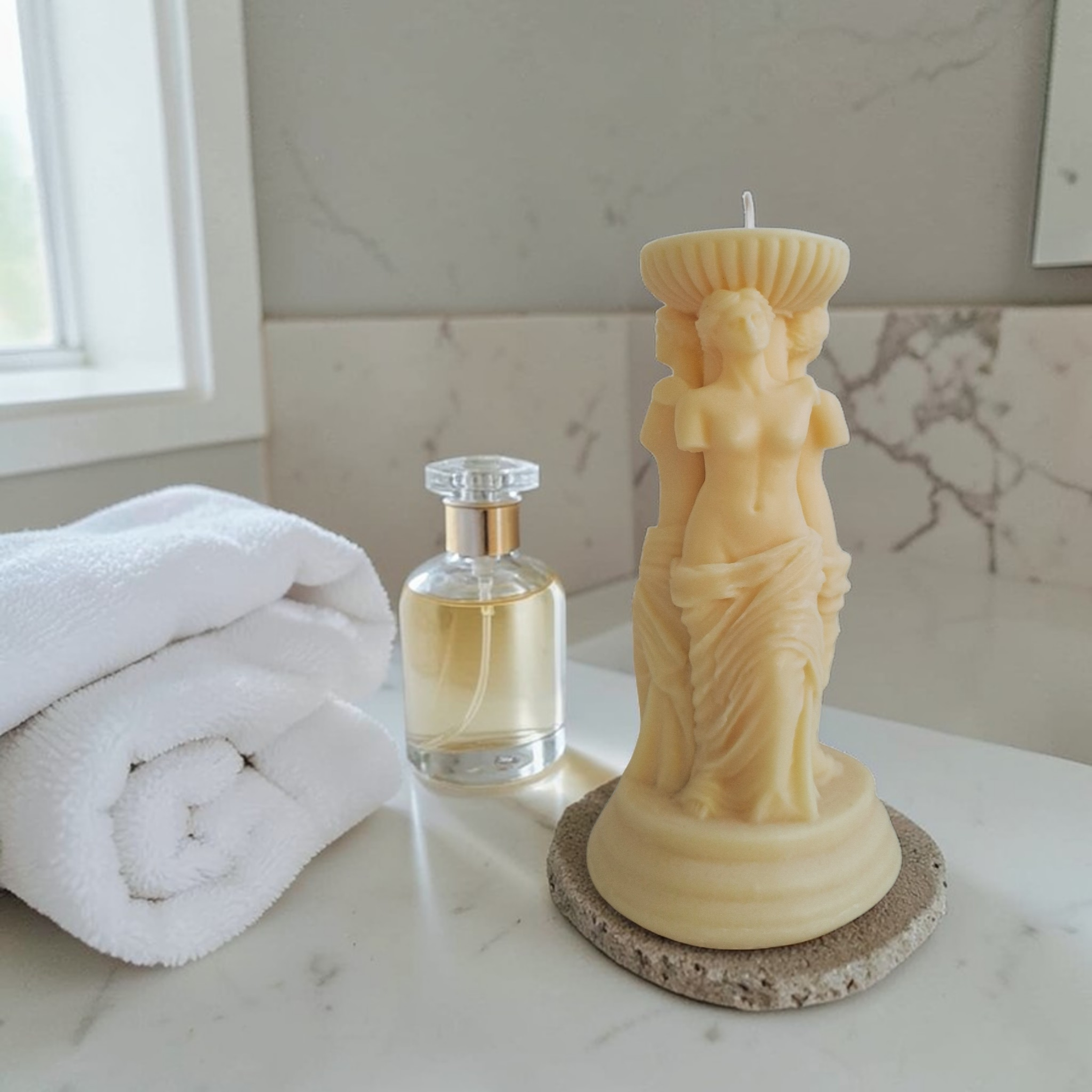 Soy wax candle shaped like a statue, of Venus , a perfume bottle, and folded towels on a marble surface.