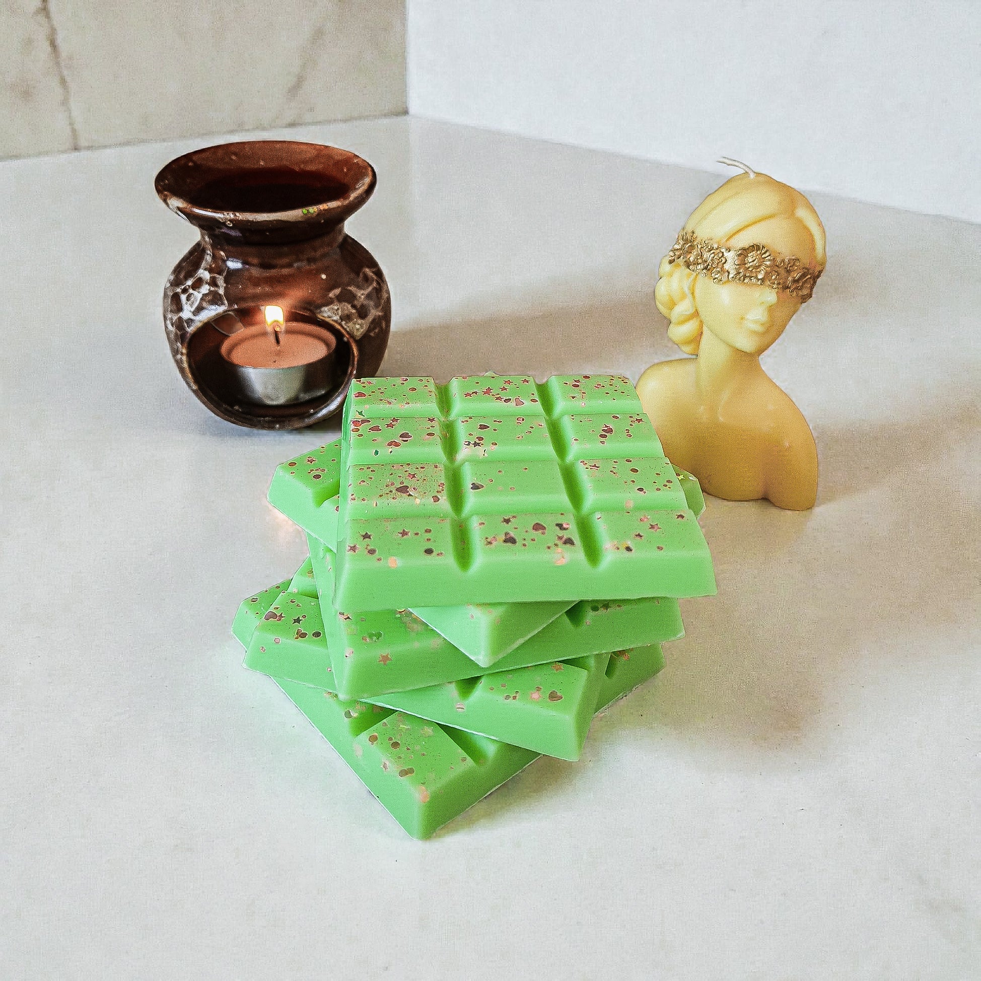 Stacked green soy wax snap bars with gold specks beside a lit candle and decorative figure on a white surface.