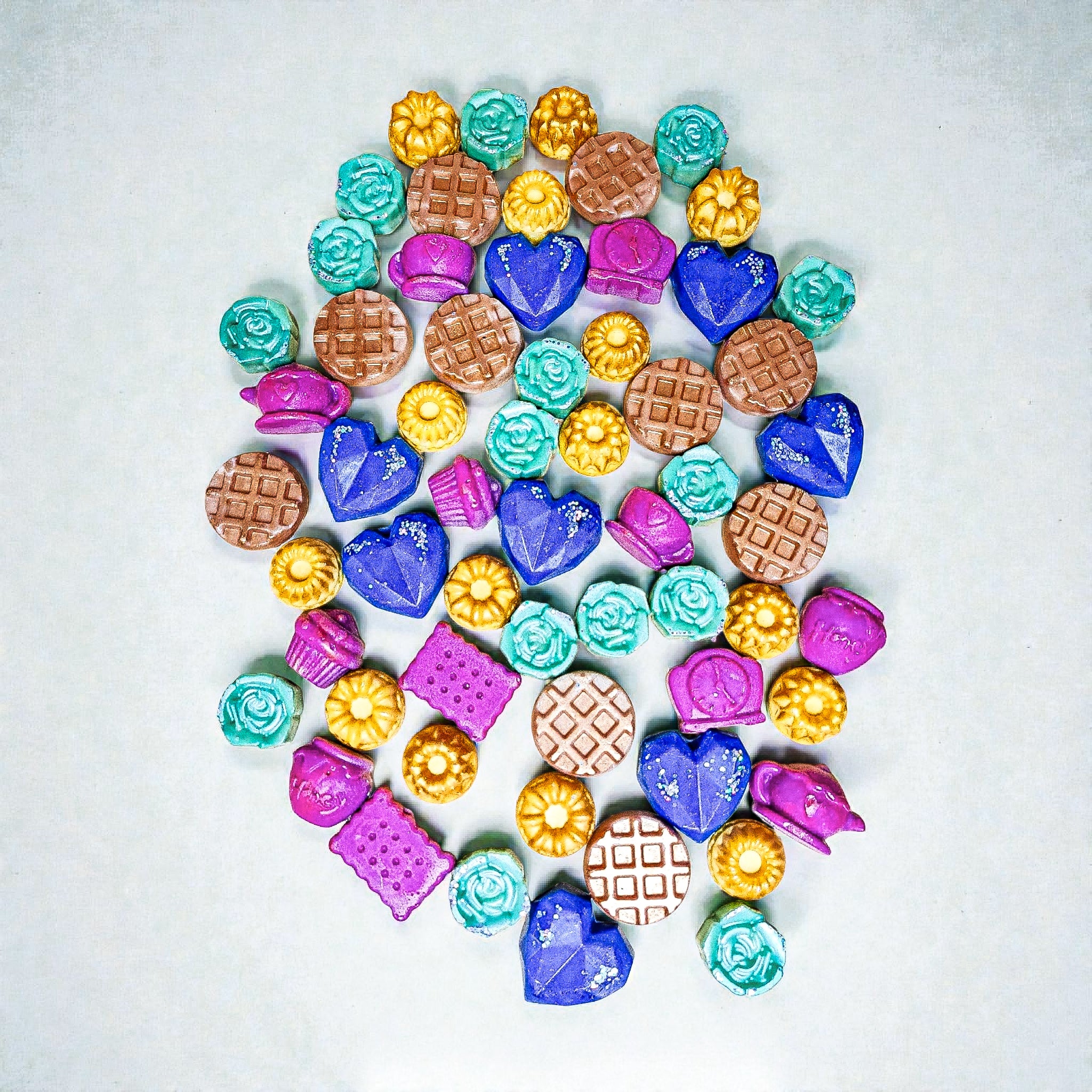 Assorted colorful soy wax melts in heart, flower and waffle shapes on a white background.