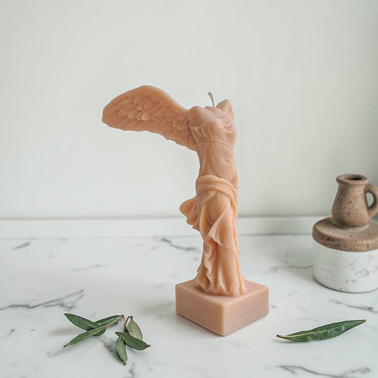 Soy wax candle shaped like a winged figure statue on a marble surface with leaves with a white background aka winged victory of Samothrace