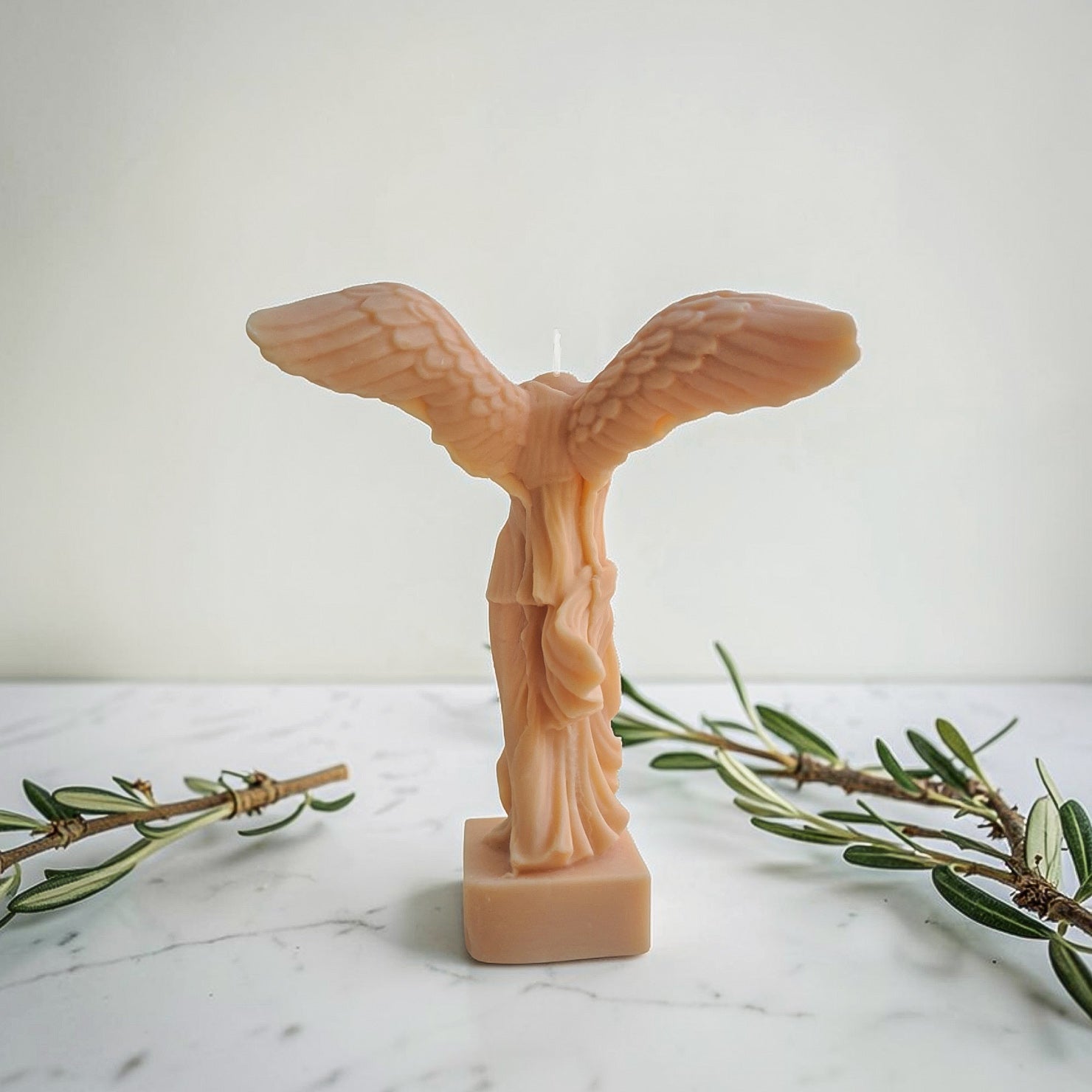 Soy wax candle shaped like a winged figure statue on a marble surface with olive branches aka winged victory of Samothrace